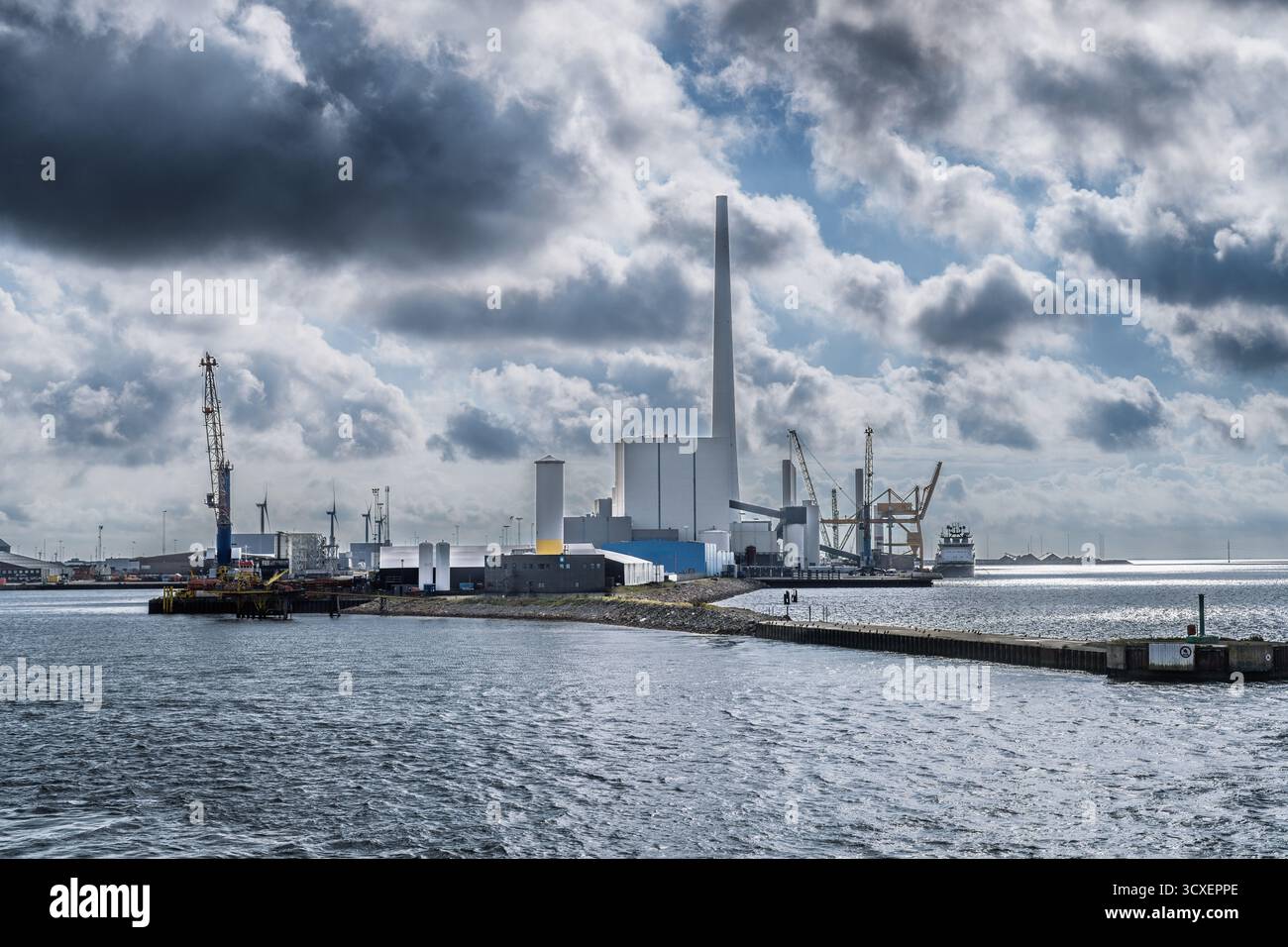 Un vivace porto industriale caratterizzato da una grande fabbrica con un'alta cisterna, gru, banchine e una nave lungo un tranquillo porto sotto le nuvole spettacolari Foto Stock