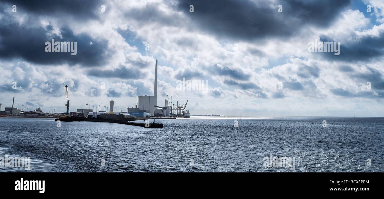 Un vivace porto industriale caratterizzato da una grande fabbrica con un'alta cisterna, gru, banchine e una nave lungo un tranquillo porto sotto le nuvole spettacolari Foto Stock