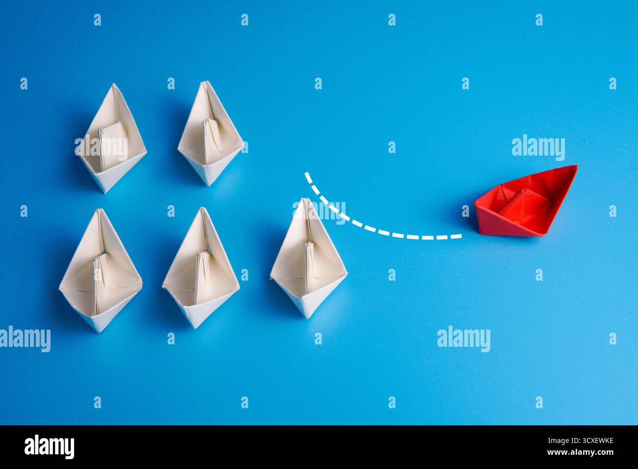Barca di carta rossa che cambia direzione allontanandosi da un gruppo di barche bianche con linee tratteggiate su sfondo blu, simboleggiando innovazione e coraggio. Foto Stock