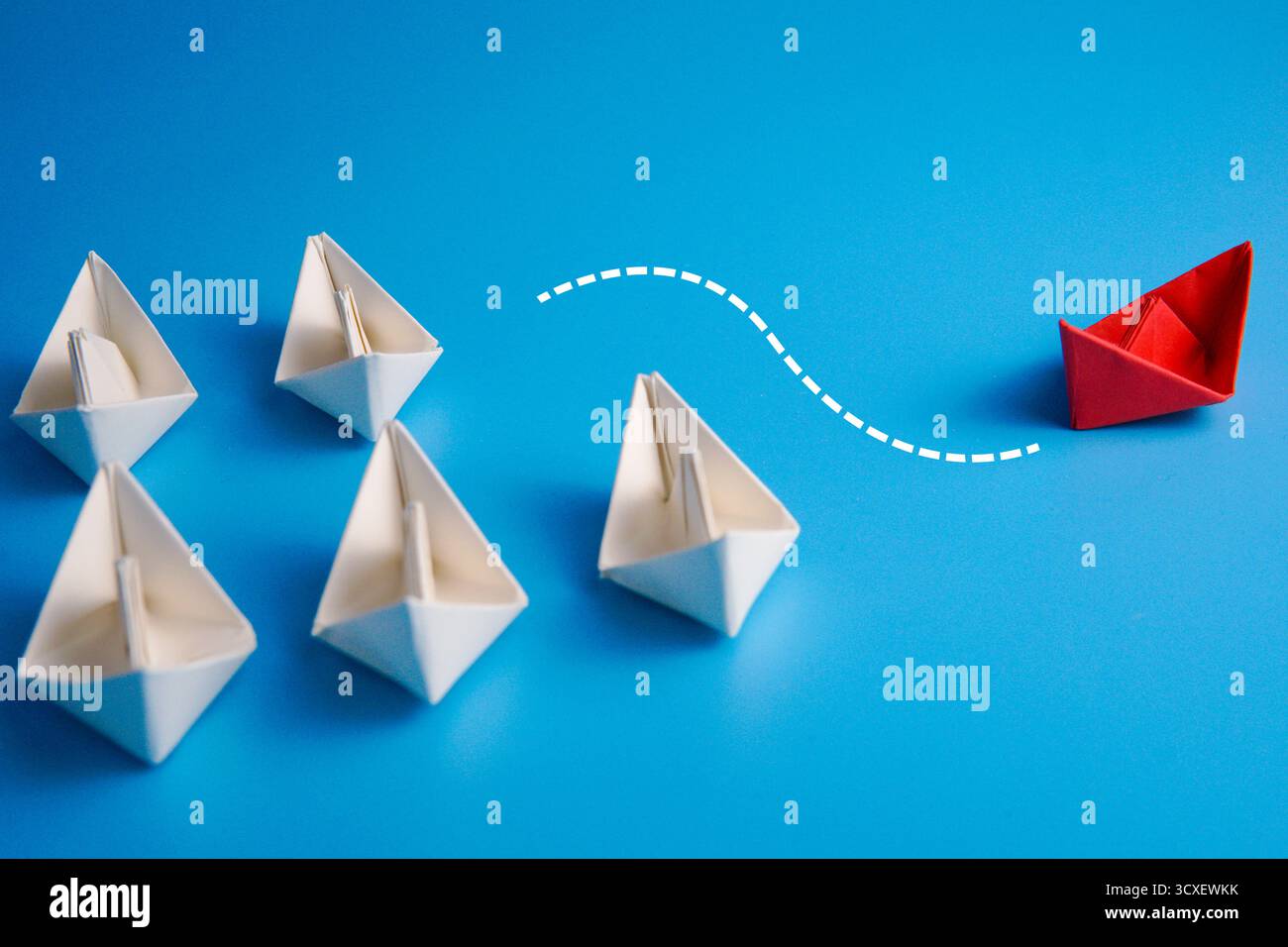 Barca di carta rossa che cambia direzione allontanandosi da un gruppo di barche bianche con linee tratteggiate su sfondo blu, simboleggiando innovazione e coraggio. Foto Stock