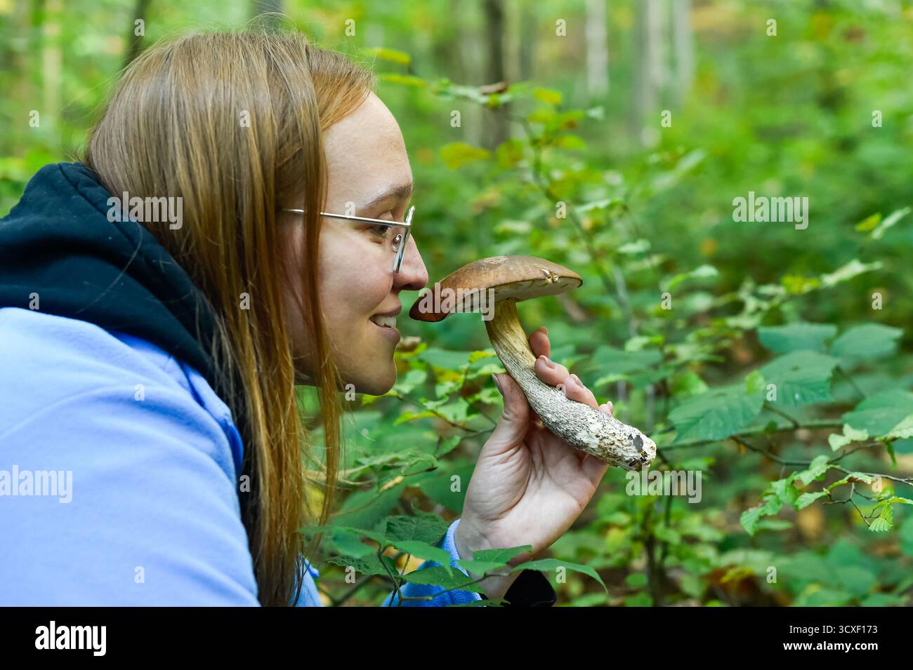 Una donna felice in bicchieri odora un fungo selvatico appena raccolto, probabilmente un Boletus, durante il foraggiamento in una verde foresta autunnale. Questa foto orizzontale cattura la gioia della caccia ai funghi. Foto Stock