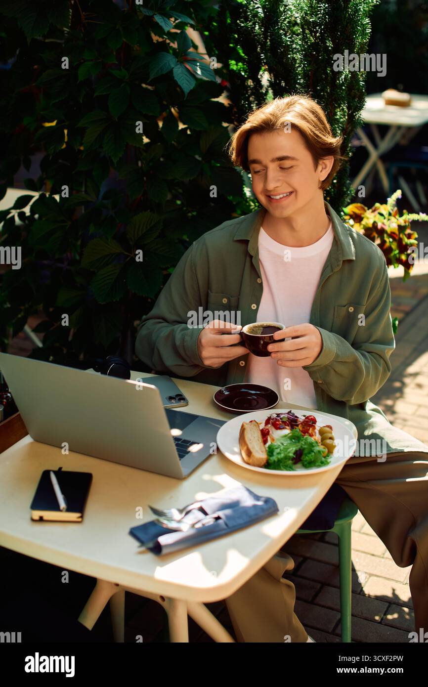 Un giovane uomo con una camicia casual lavora sul suo computer portatile mentre sorseggia un caffè in un vivace bar. Foto Stock