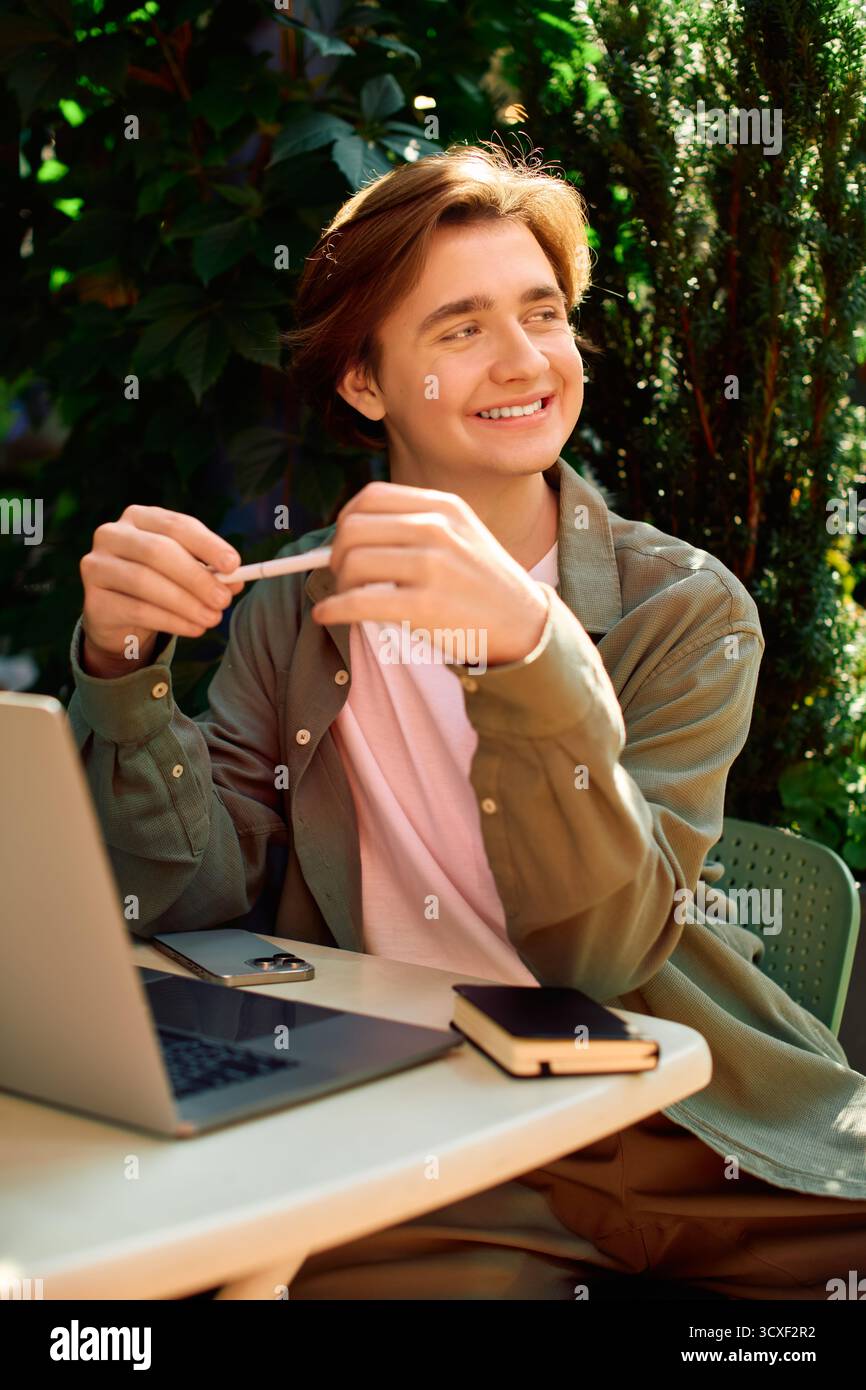 Un giovane uomo siede ad un tavolo da caffè, lavora sul suo portatile e sorride, circondato dal verde. Foto Stock