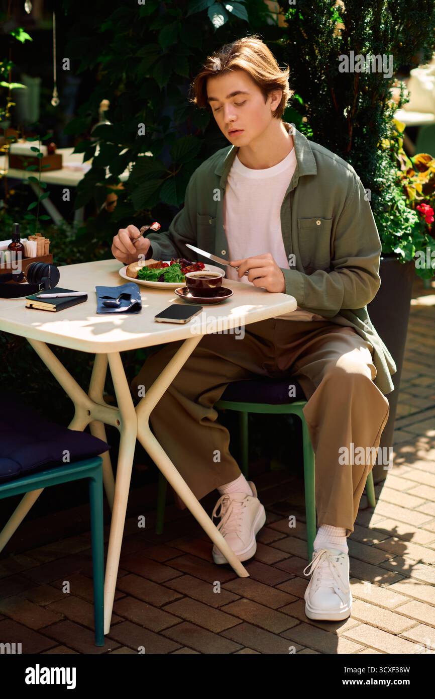 Un giovane con una camicia verde oliva si siede a un tavolo, gustando il suo pasto in un elegante caffè. Foto Stock