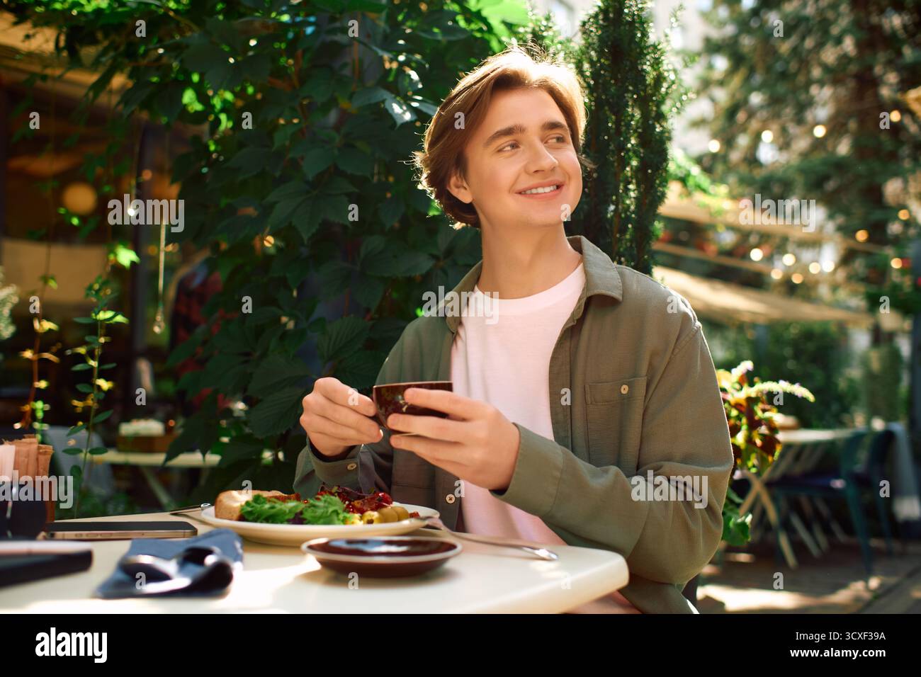 Un giovane uomo con una camicia verde oliva sta assaporando il suo pasto in un elegante caffè pieno di verde. Foto Stock