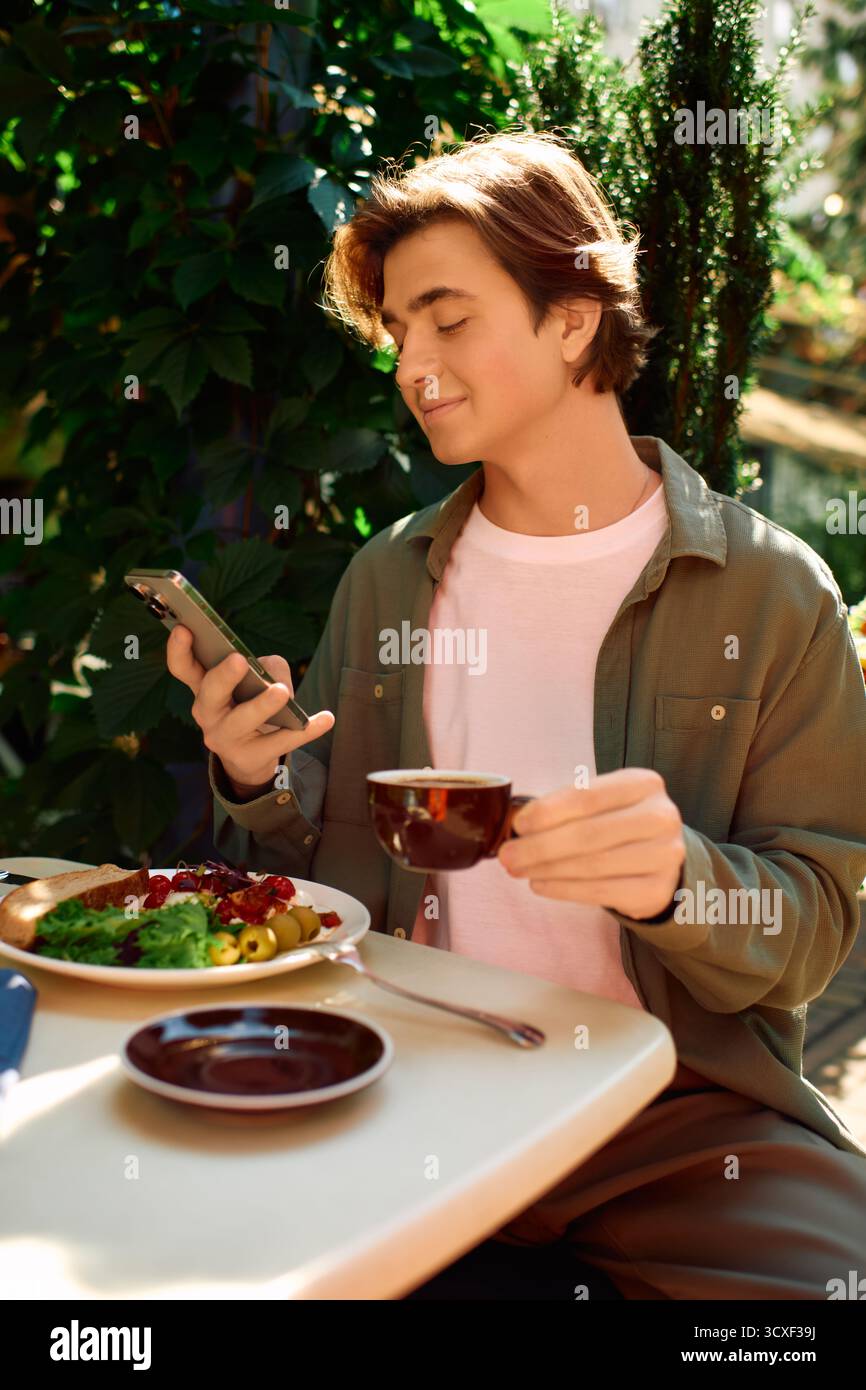 Un giovane con una camicia verde oliva si diverte a mangiare e usa il telefono in un vivace caffè. Foto Stock