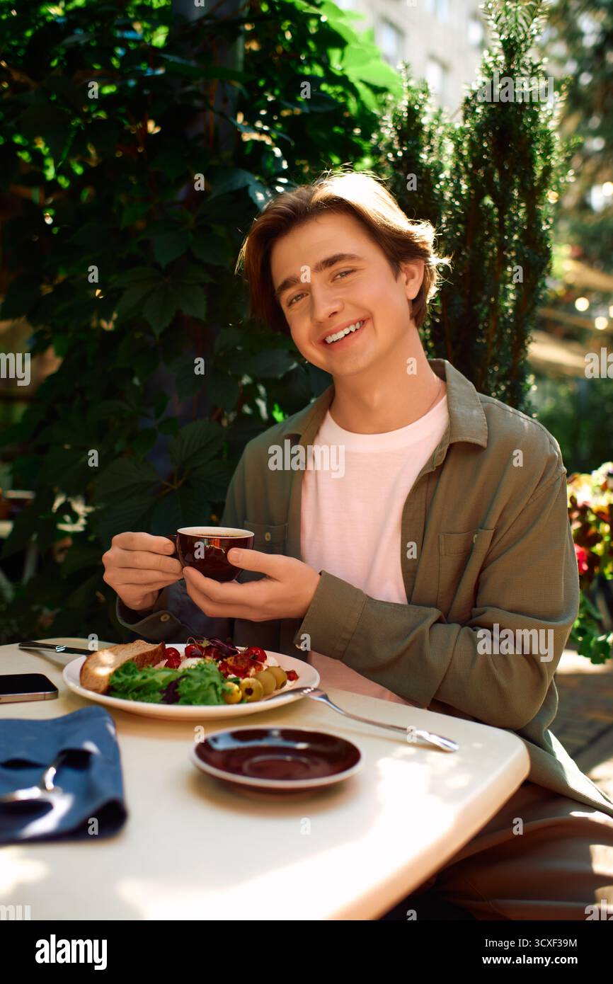Un giovane uomo con una camicia d'oliva sorride mentre si gusta il suo pasto in un bar alla moda. Foto Stock