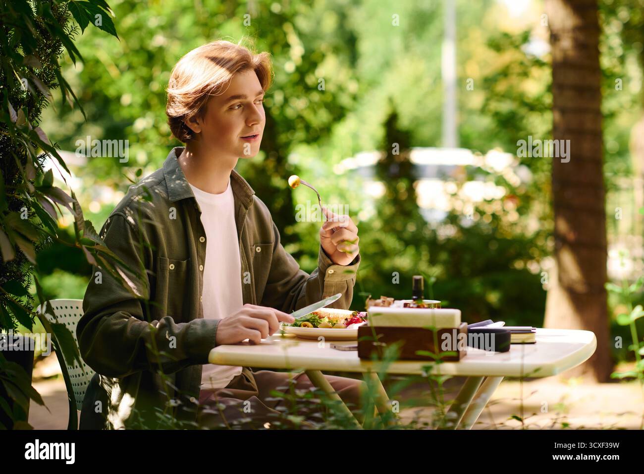 Un giovane con una camicia verde oliva si diverte a mangiare all'aperto in un vivace caffè. Foto Stock