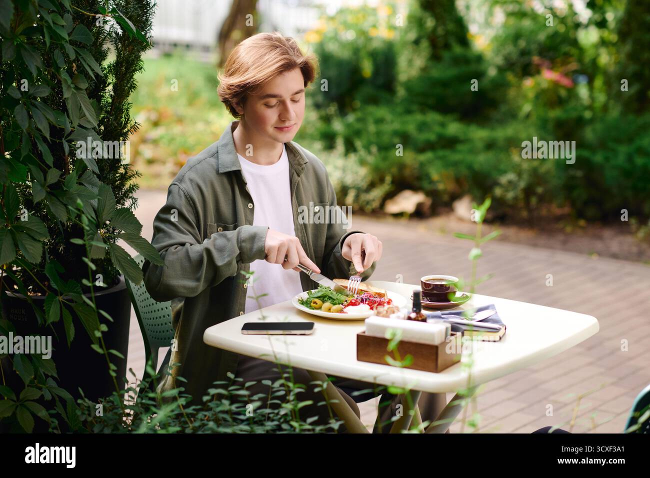 Un giovane uomo con una camicia verde oliva sta assaporando il suo pasto in un accogliente caffè circondato dalla natura. Foto Stock