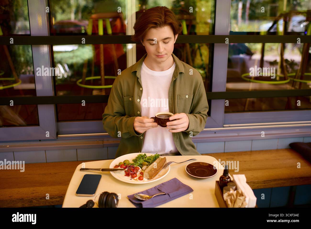 Un giovane con una camicia verde oliva siede in un elegante caffè, assaporando il suo pasto con un caffè. Foto Stock