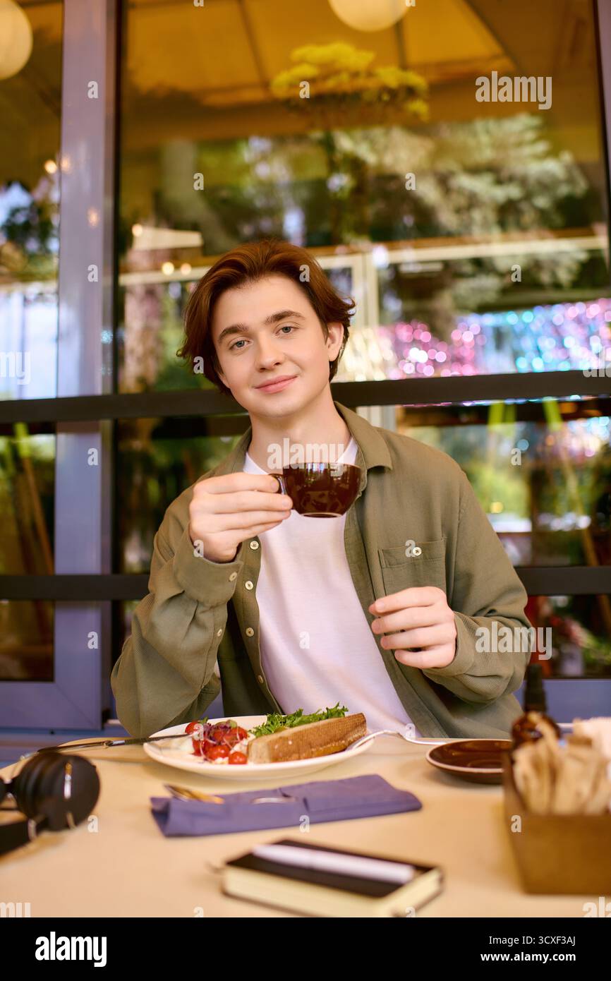 Un giovane con una camicia verde oliva siede in un bar moderno gustando il suo pasto e la sua bevanda. Foto Stock