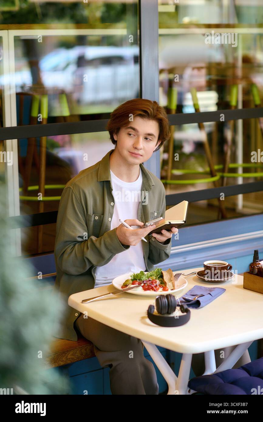 In un elegante caffè, un giovane con una camicia verde oliva gusta il suo pasto mentre legge. Foto Stock