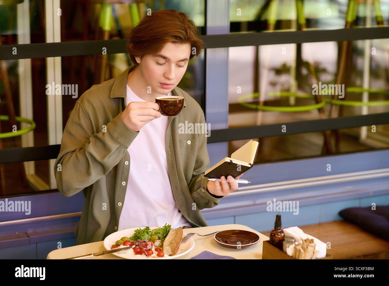 Un giovane uomo con una camicia verde oliva sta assaporando il suo pasto e il caffè mentre tira giù le note. Foto Stock