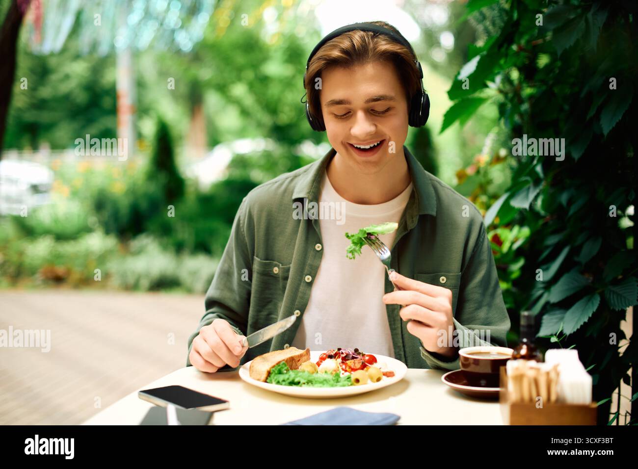 Un giovane con una camicia verde oliva mangia felicemente il suo pasto mentre si rilassa in un bar. Foto Stock