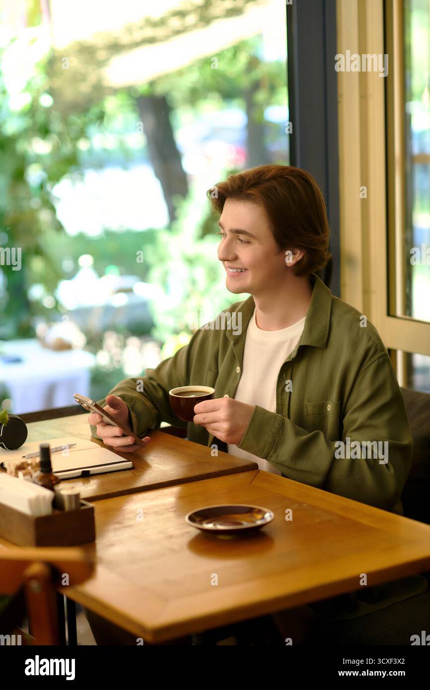 Un giovane con una camicia è concentrato sul suo smartphone mentre sorseggia un caffè in un bar moderno. Foto Stock