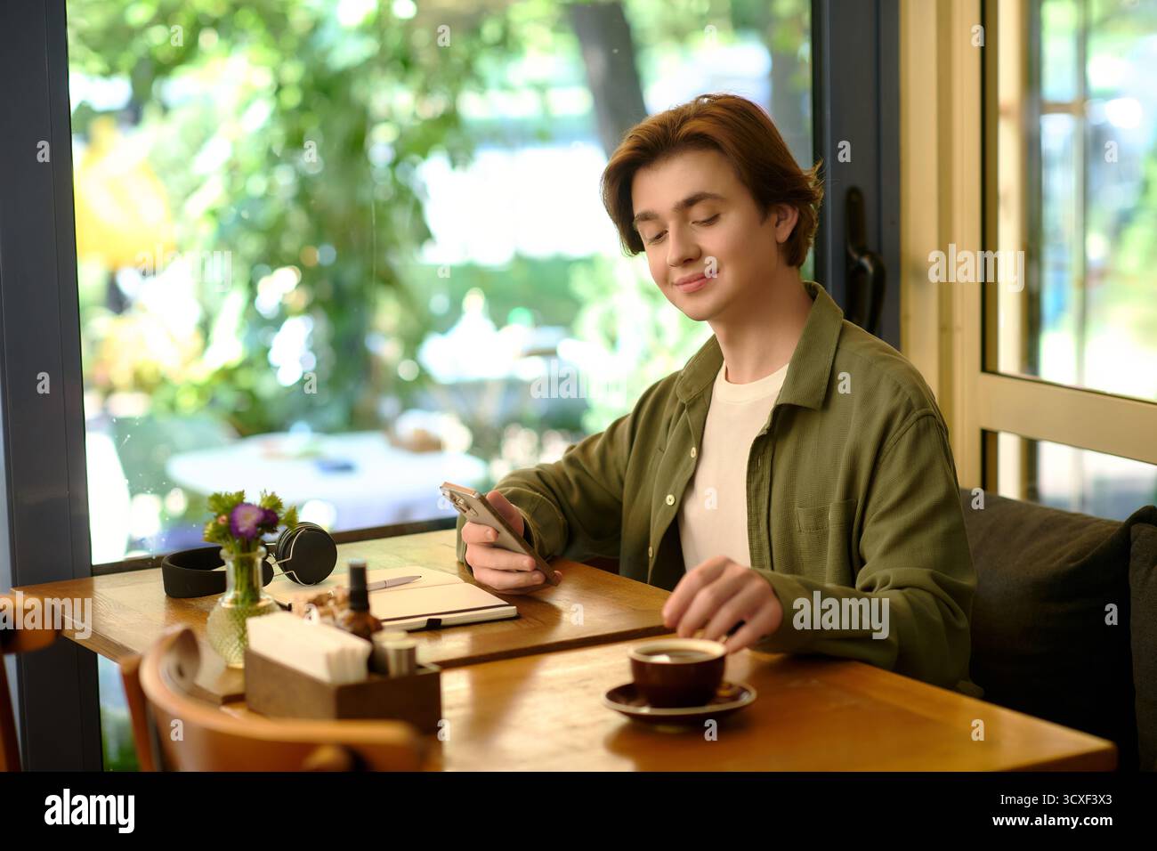 Un giovane con una camicia si concentra sul suo smartphone in un bar alla moda, sorseggiando un caffè e lavorando. Foto Stock