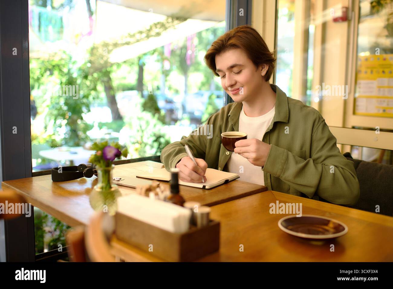 Un giovane uomo in camicia scrive nel suo taccuino e sorseggia un caffè in un vivace caffè. Foto Stock