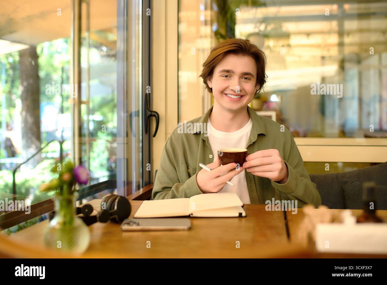 Un giovane uomo siede in un caffè moderno, sorridendo mentre tiene una tazza di caffè Foto Stock