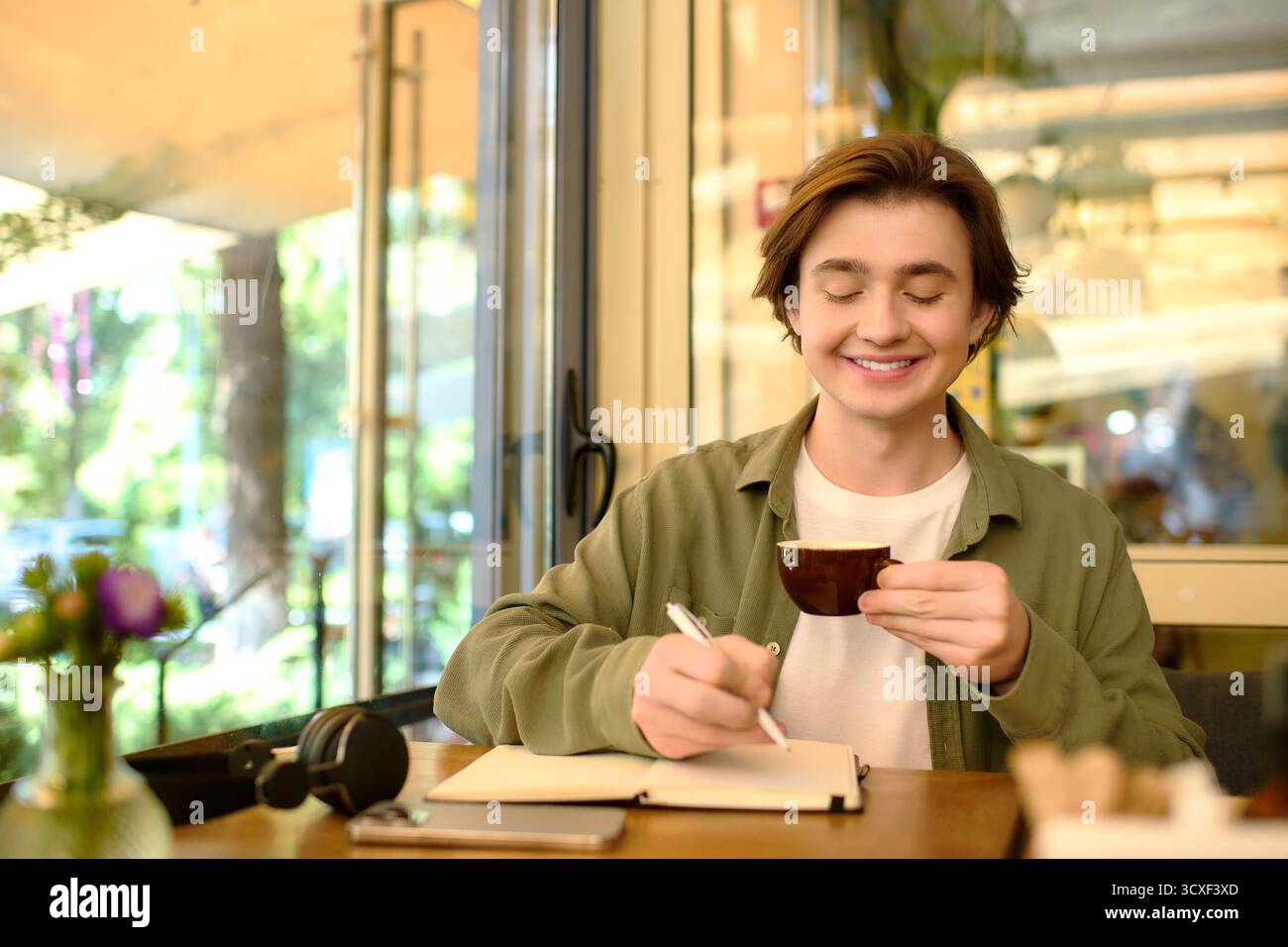 Un giovane in camicia si diverte a prendere un caffè e prendere appunti in un caffè alla moda. Foto Stock
