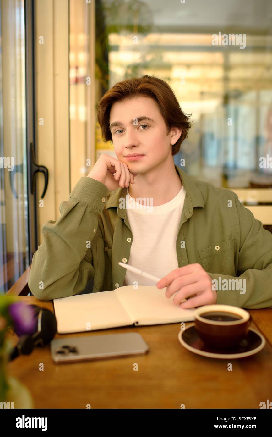 Un giovane uomo in camicia si concentra sulle sue idee in un caffè contemporaneo, mescolando studio e tempo libero. Foto Stock