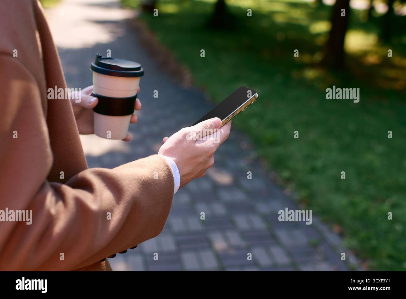 Un giovane con un cappotto elegante ama il caffè all'aperto in un parco autunnale, controllando il telefono. Foto Stock