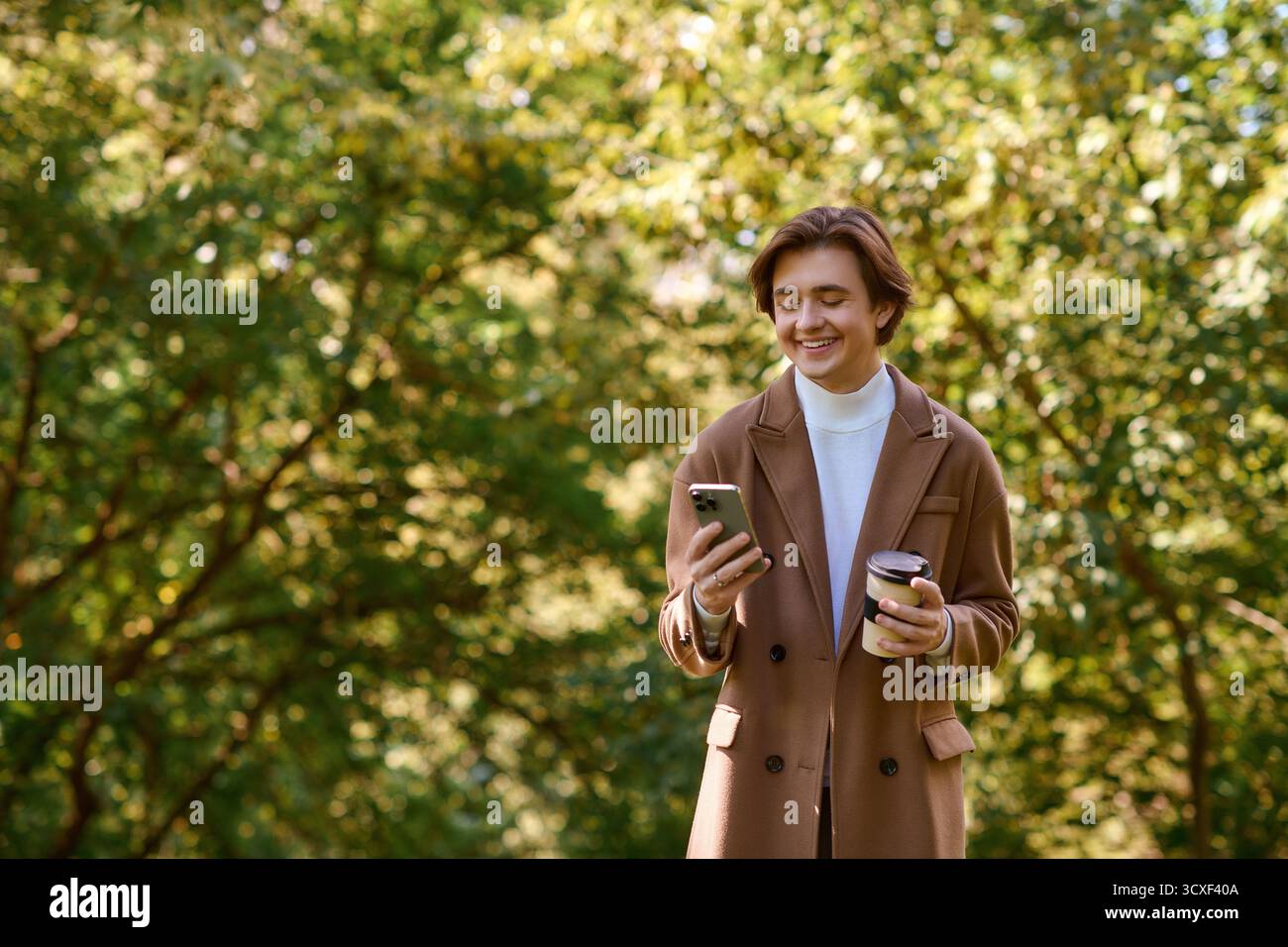 In un parco autunnale, un giovane con un cappotto elegante sorride mentre usa il telefono e tiene un drink. Foto Stock