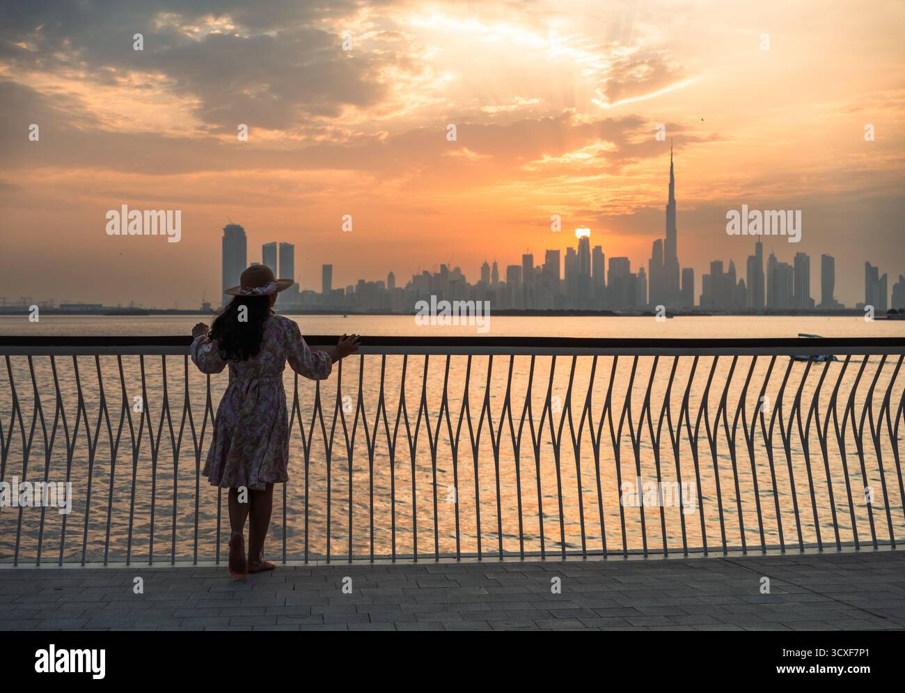 La silhouette di una donna si erge su un molo vicino al mare al tramonto, guardando la vista dello skyline di Dubai Foto Stock
