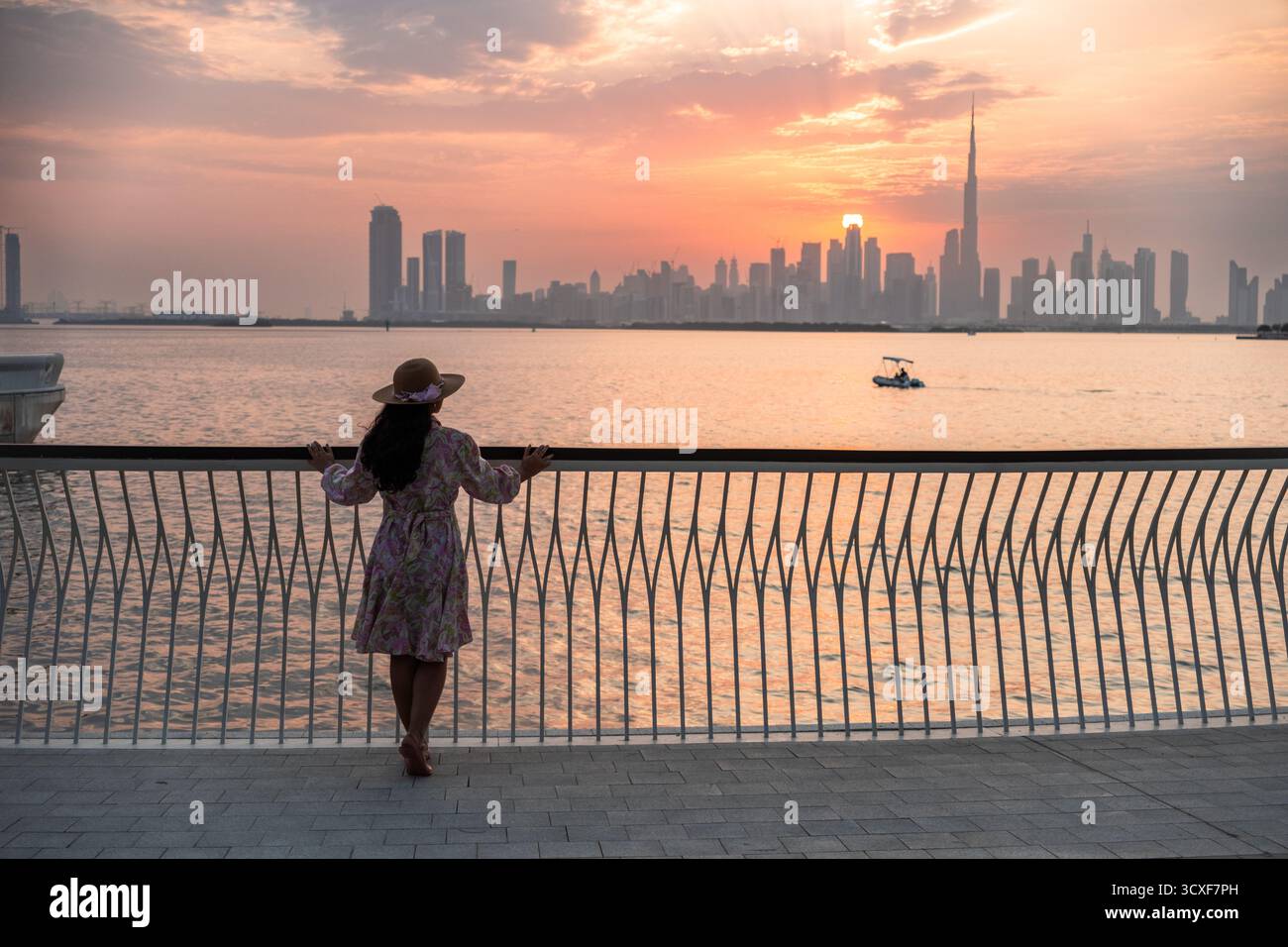 La silhouette di una donna che indossa un cappello si erge su un molo vicino al mare al tramonto, guardando la vista dello skyline di Dubai Foto Stock