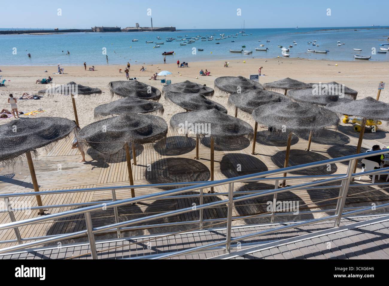 Vista estiva della spiaggia di Cadice, Andalusia, Spagna, con ombrelloni colorati e barche ormeggiate sulle calme acque marine. Tipica scena di tou sul mare spagnolo Foto Stock