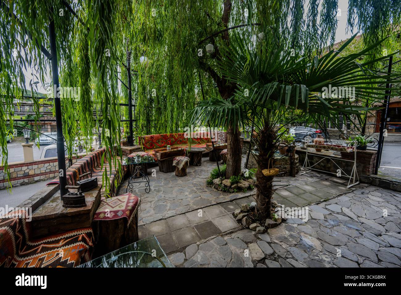 Area salotto all'aperto ombreggiata di un tradizionale ristorante azero a Sheki, caratterizzato da pavimento in pietra, mobili rustici e vegetazione lussureggiante. Foto Stock