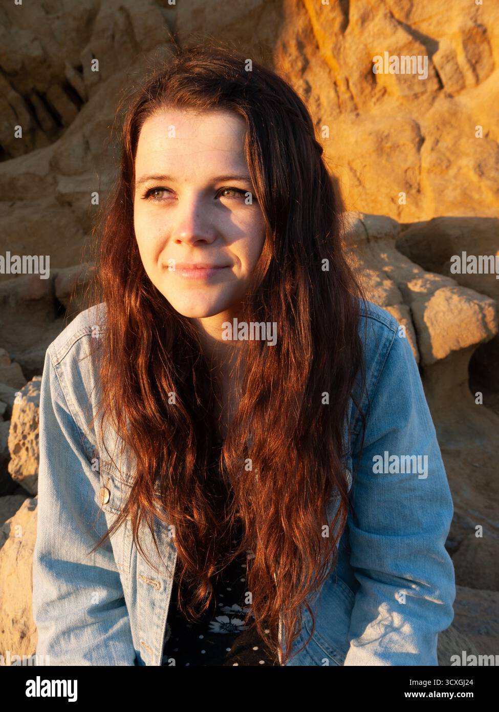 Giovane donna con lunghi capelli auburn in giacca di denim seduta sulle rocce costiere sotto la luce dorata della sera Foto Stock