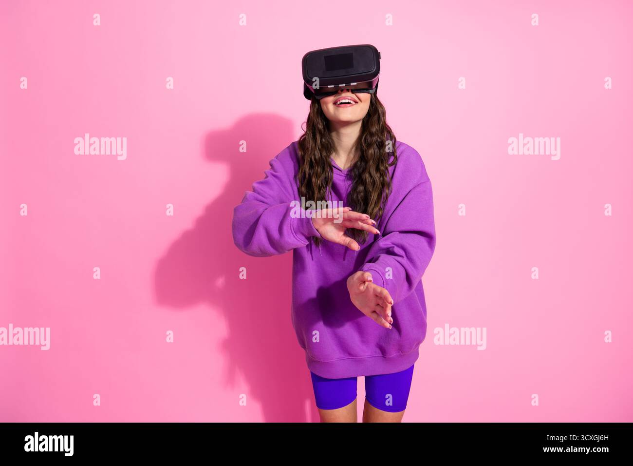 Giovane donna in un abbigliamento sportivo alla moda che utilizza il visore per la realtà virtuale su un vivace sfondo rosa Foto Stock
