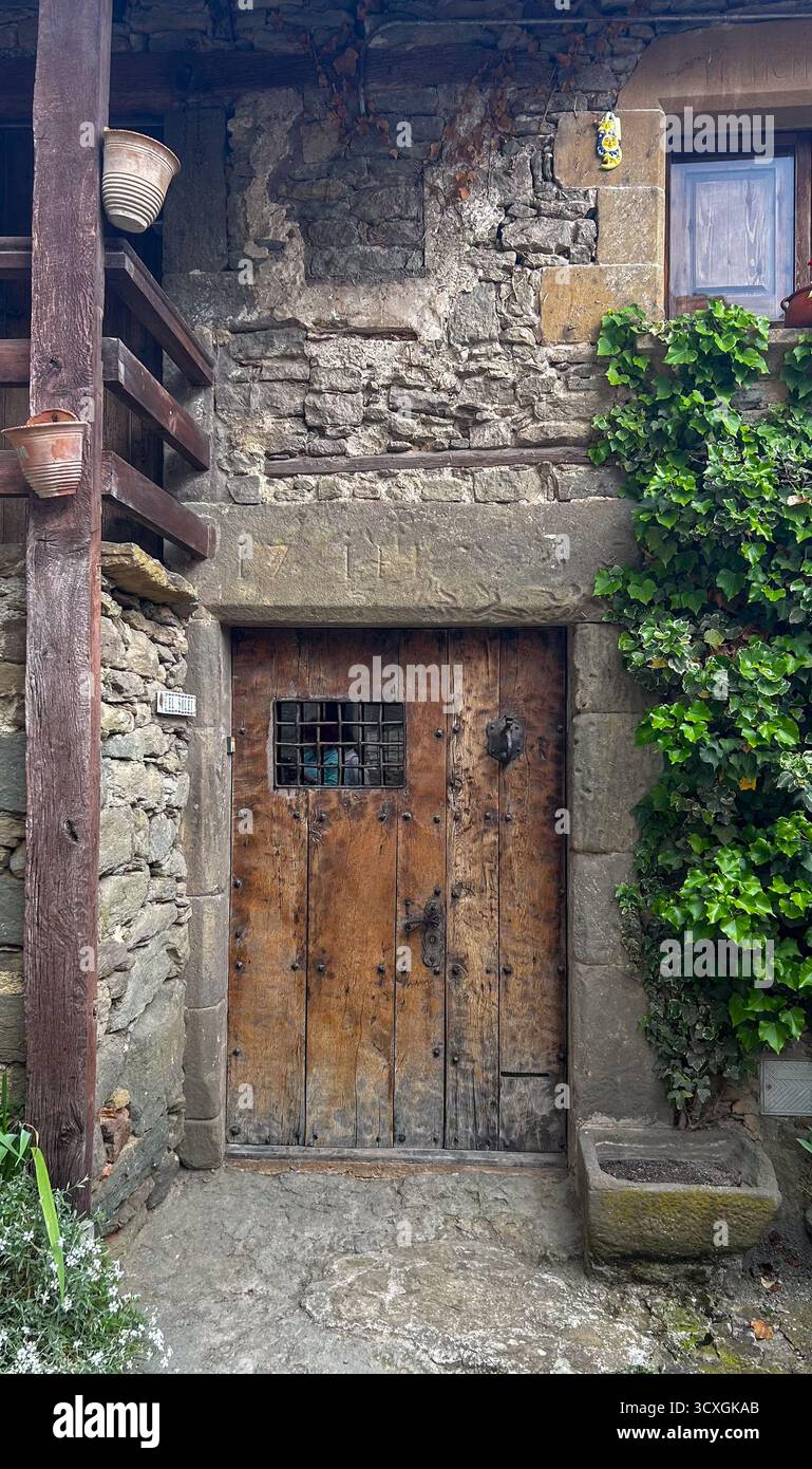 Porta in legno d'epoca con griglia in metallo incastonata in una vecchia parete in pietra di una casa rustica. L'arrampicata sull'edera sulle pareti e le texture invecchiate creano un'atmosfera storica Foto Stock