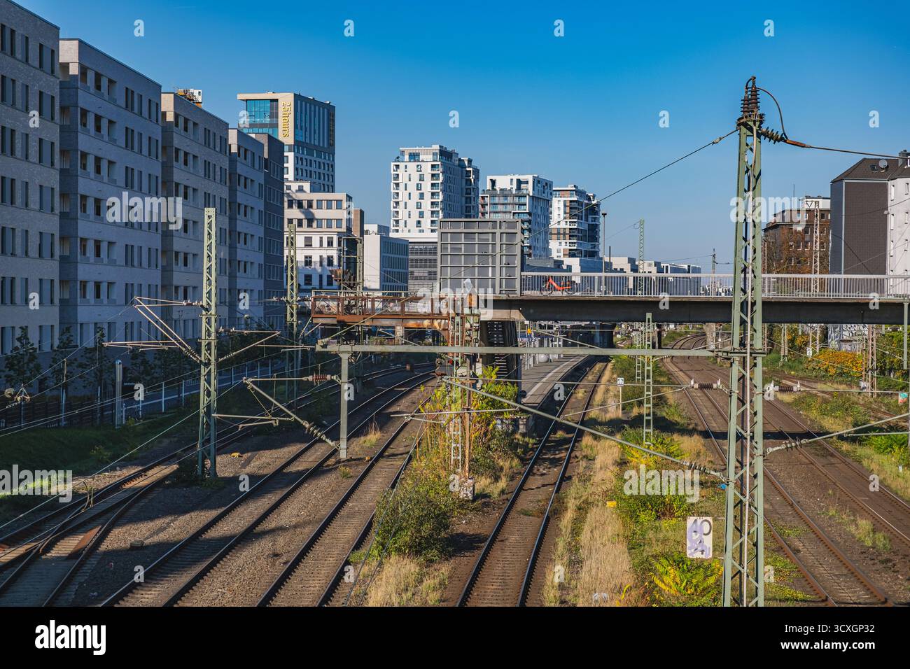 Vista dei binari ferroviari vicino alla stazione ferroviaria centrale incorniciati da moderni edifici residenziali e da un ponte pedonale sotto un cielo azzurro Foto Stock