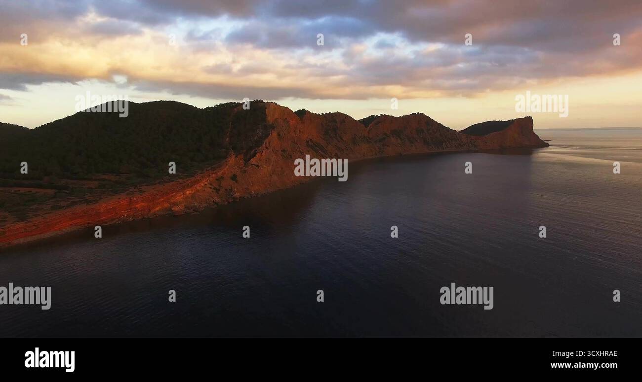 Con scogliere rocciose costiere che si innalzano dall'oceano calmo sotto il cielo pastello con nuvole e creste boscose Foto Stock