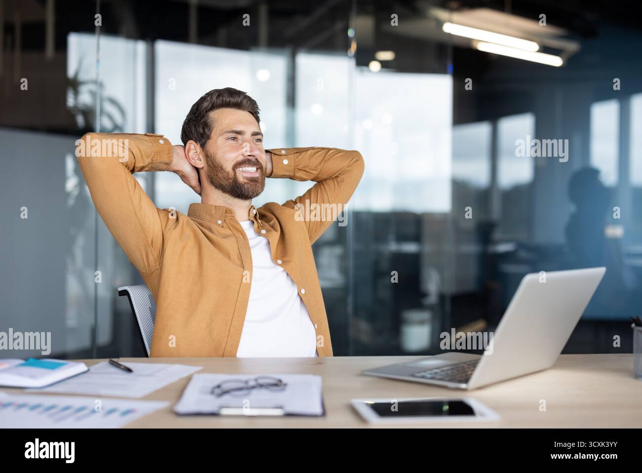 Uomo d'affari felice che si gode una pausa rilassante alla scrivania dell'ufficio, si piega indietro con le mani dietro la testa, sorride e sembra soddisfatto del suo lavoro a. Foto Stock