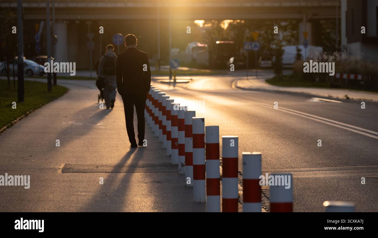 Scena urbana al tramonto con un uomo che cammina e una persona con un passeggino lungo una strada costeggiata da pali di sicurezza rossi e bianchi. Luce calda della sera e lon Foto Stock Scena urbana al tramonto con un uomo che cammina e una persona con un passeggino lungo una strada costeggiata da pali di sicurezza rossi e bianchi. Luce calda della sera e lon Foto Stock