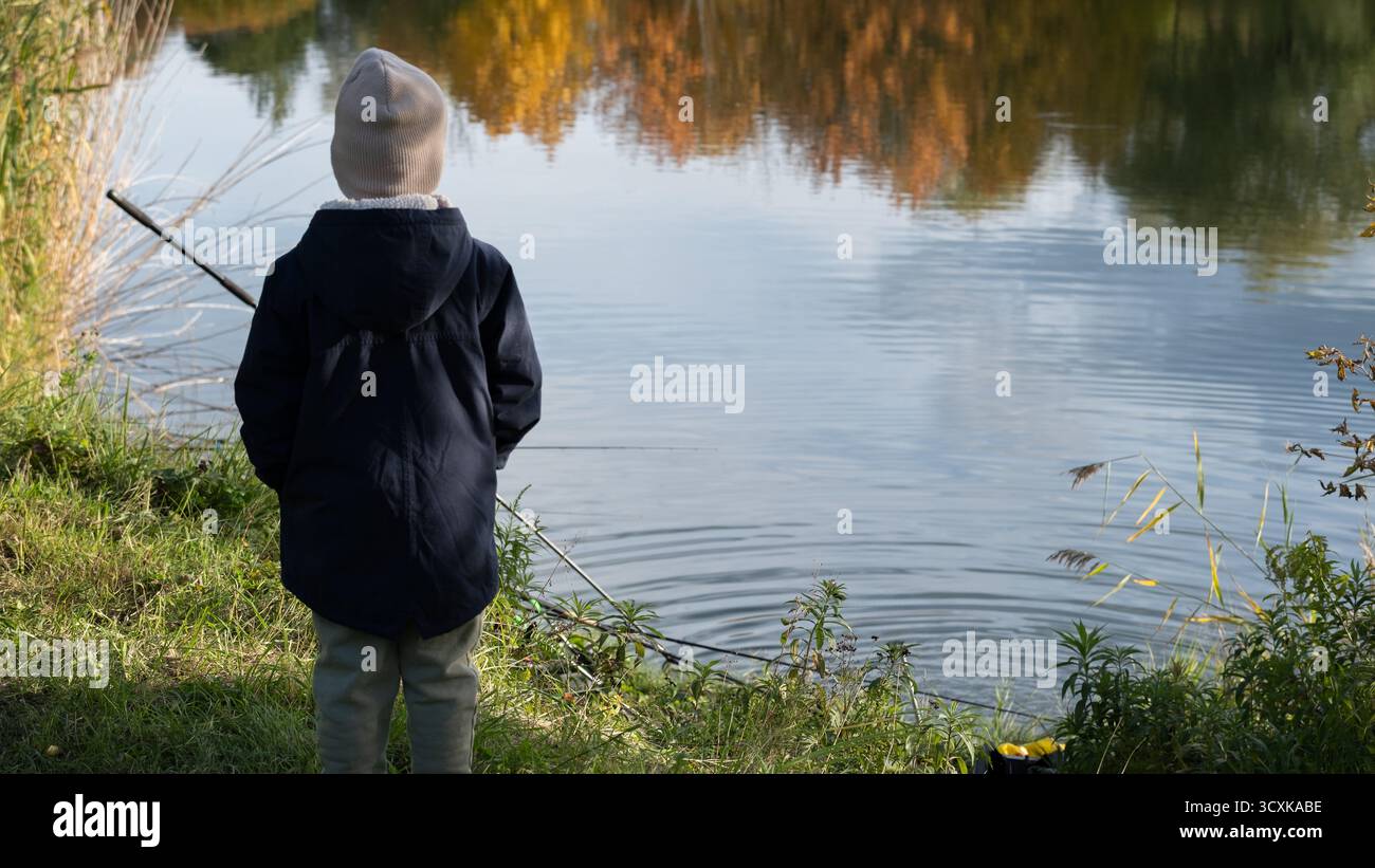 Foto realistica di un bambino in abiti caldi in piedi vicino a un tranquillo lago autunnale con una canna da pesca. Autentica scena all'aperto che rappresenta i curiosi dell'infanzia Foto Stock