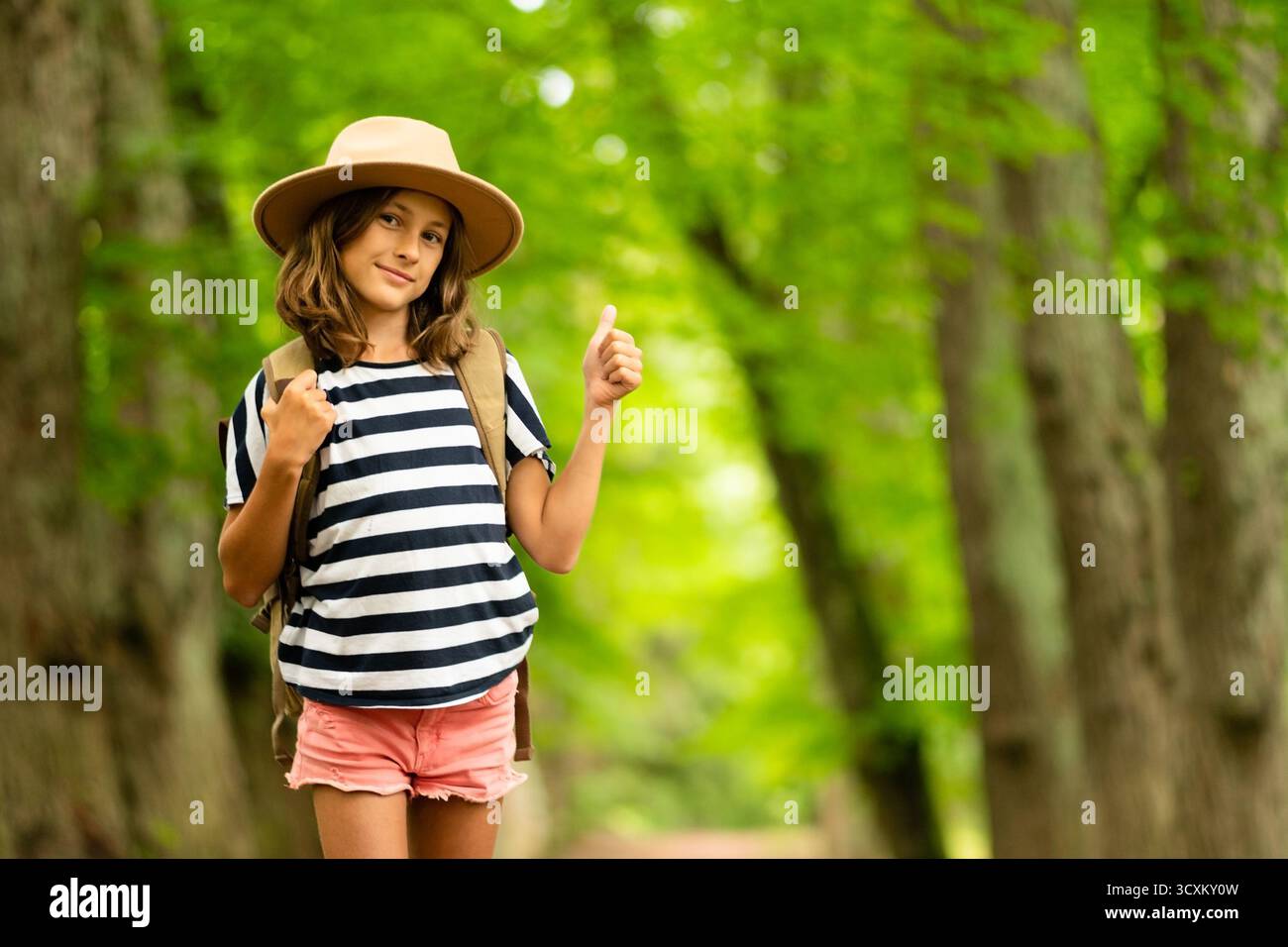Ragazza radiosa con attrezzatura da trekking sorride brillantemente alla macchina fotografica, in piedi sul sentiero della foresta. Incarna lo spirito d'avventura estivo giovanile. Foto Stock