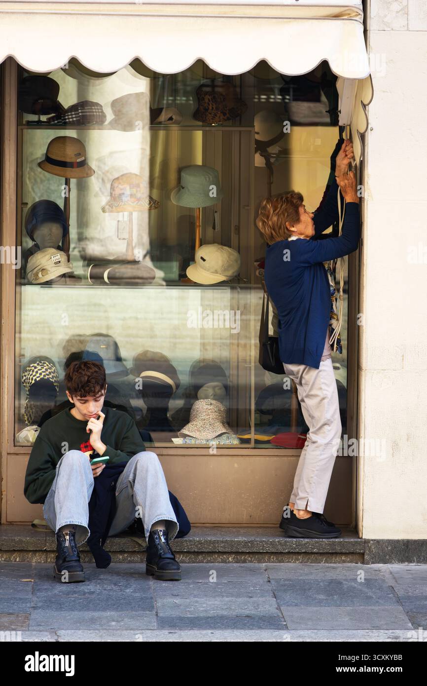 Un giovane uomo si siede premurosamente vicino a un negozio di cappelli, mentre una donna più anziana adatta la tenda in una strada tranquilla. Foto Stock