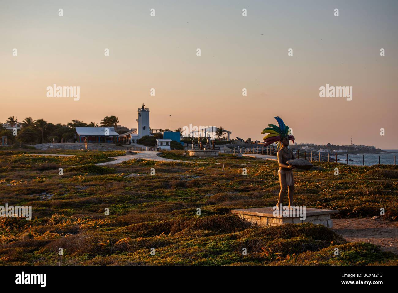 Isla Mujeres, Cancun, Messico Statua di un uomo che indossa un copricapo colorato si trova in un campo vicino a una spiaggia. Il cielo è un bel mix di arancione e blu Foto Stock