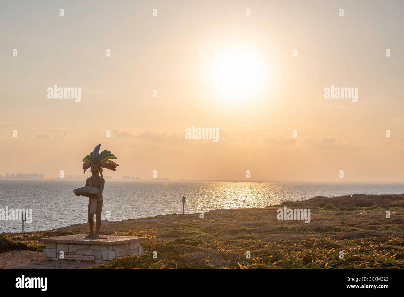 Isla Mujeres, Cancun, Messico Statua di una persona che indossa un copricapo di piume si erge su una collina che si affaccia sull'oceano. Il cielo è un mix di arancione e blu Foto Stock