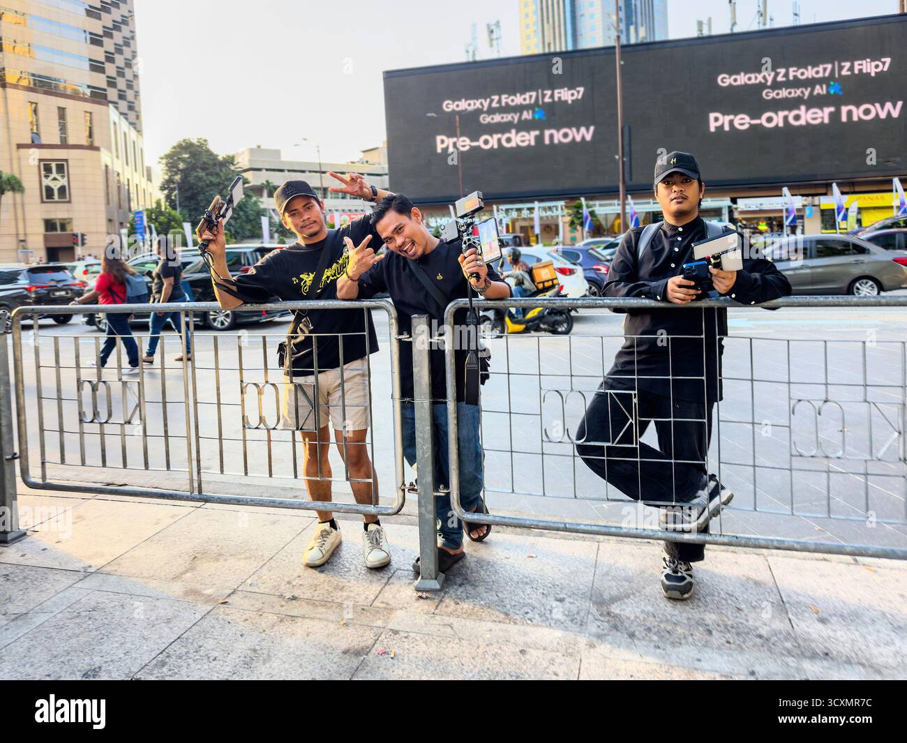 Tre giovani uomini asiatici con attrezzatura fotografica che scattano foto turistiche di fronte a un grande cartellone digitale vicino alle vivaci torri petronas di kuala lumpur - Immagine stock catturata con smartphone