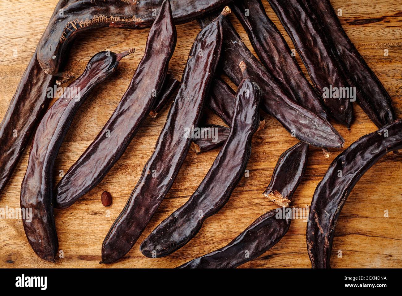 un gruppo di cialde di carrubo maturo si estende su una superficie in legno, catturati in dettaglio per evidenziare la loro forma allungata e le ricche tonalità di marrone scuro Foto Stock