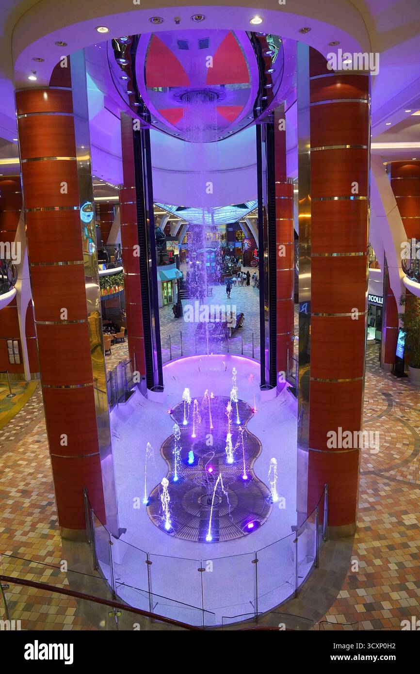Fontana illuminata e soffitto a cascata sono a bordo di Allure of the Seas, illuminata di luce viola e blu all'interno della Royal Promenade della nave Foto Stock