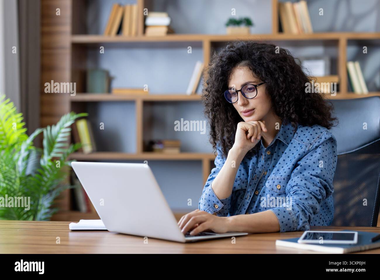 Giovane donna con capelli ricci e occhiali che si concentrano mentre digita su un computer portatile su una scrivania di legno in un moderno ufficio domestico, simboleggiando il lavoro remoto e la p Foto Stock