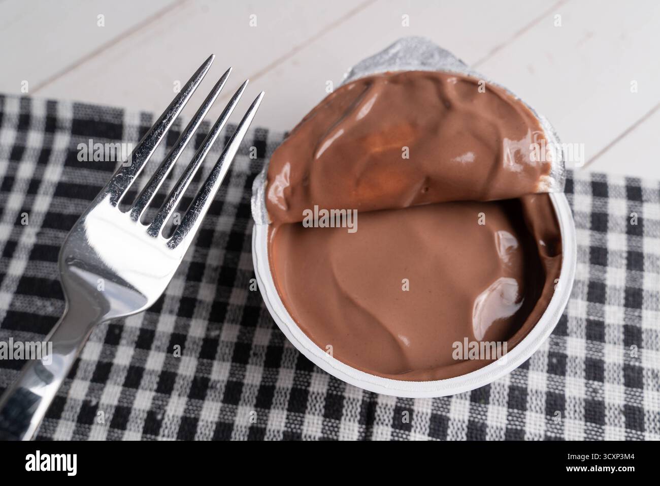 Baviera, Germania - 15 ottobre 2025: La tendenza a mangiare pudding con una forchetta si sta diffondendo rapidamente sui social media e sta scatenando un dibattito. Accanto a una forchetta si trova una tazza aperta di budino al cioccolato che simboleggia una bizzarra tendenza alimentare. *** Der Trend, Pudding mit einer Gabel zu essen, verbreitet sich rasant in sozialen Medien und sorgt für Diskussionen. Ein offener Schokopuddingbecher liegt neben einer Gabel Sinnbild eines skurrilen Foodtrends. Foto Stock