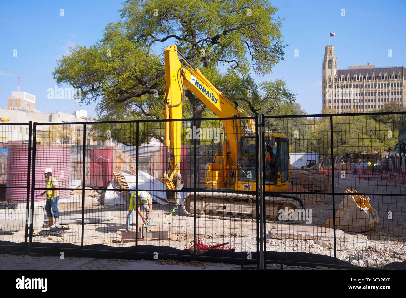 I membri dell'equipaggio lavorano in un cantiere recintato con un escavatore Komatsu giallo vicino ad Alamo Plaza, mentre la bandiera del Texas ondeggia in cima a un edificio vicino Foto Stock
