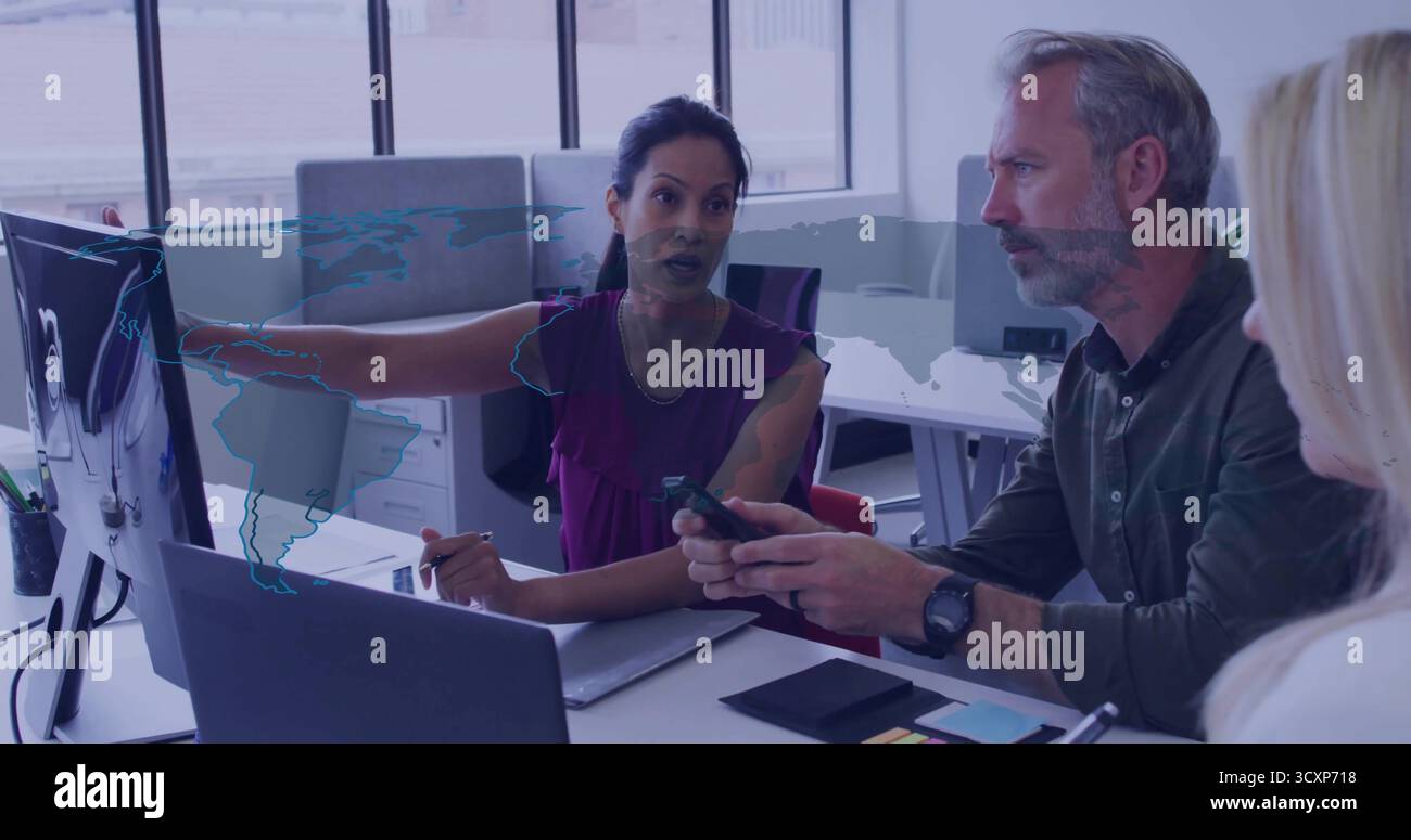 Donna dai capelli scuri che punta il monitor del computer in ufficio, con laptop e smartphone Foto Stock