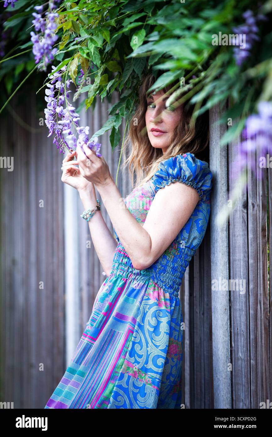 Bella signora con un abito estivo a motivi blu, che si rilassa per recinzione sotto un cespuglio di wysteria, occhi che guardano verso la macchina fotografica, accarezzano e odorano fiori Foto Stock